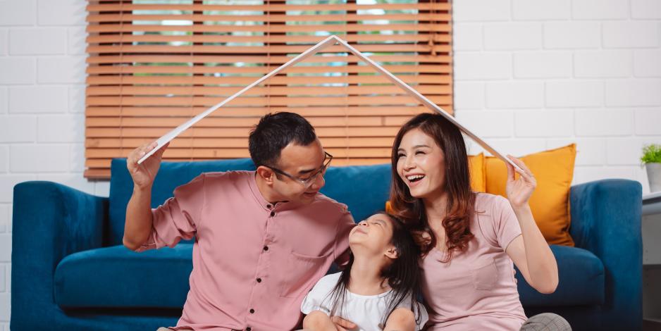 Happy family holding a model roof over their head as they sit and laugh in a living room